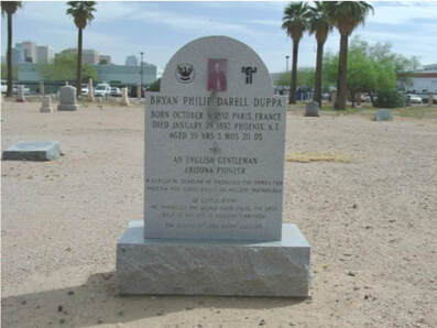 A snapshot of an upright gravestone set in a stone base in a desert cemetery with palm trees in the background; text includes “Bryan Philip Darell Duppa; birth and death dates; An English Gentleman; Arizona Pioneer”