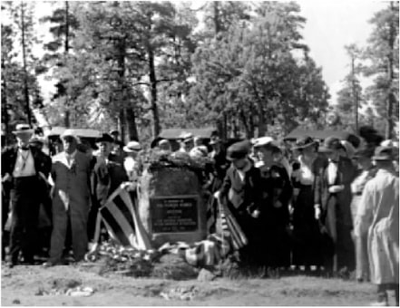 A photograph of a group of people dressed in early 20th century clothing gathered in front of a stone about 5 feet tall with a marker.