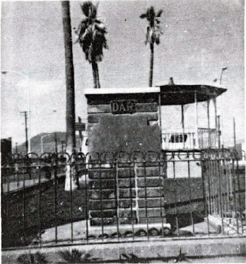 A vintage snapshot of a brick marker base with “DAR” at the top and a decorative iron fence on at least two sides in a parklike setting.