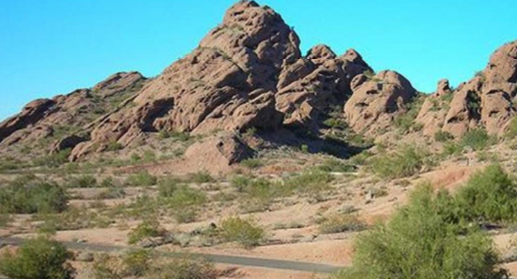 A snapshot of a Sonoran Desert scene of a reddish rock butte rising at a 45-degree angle identified as Barnes Butte. Shrubs and a narrow paved walkway in the foreground.