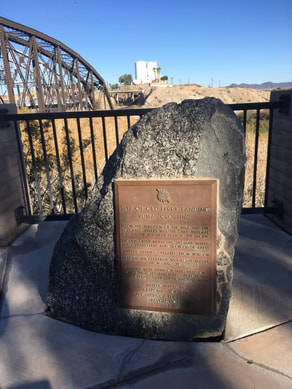 A snapshot of the large stone marker on a platform with an iron railing. A portion of an iron bridge is in the background. Marker text is in the body of this page.