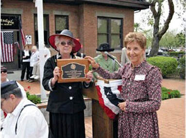 A snapshot of adults in period costume in front of the school. Two women are holding the marker.