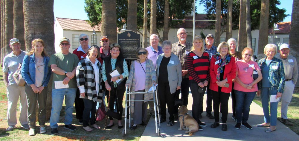 A snapshot of about eighteen people and one dog gathered in front of the school building and marker.