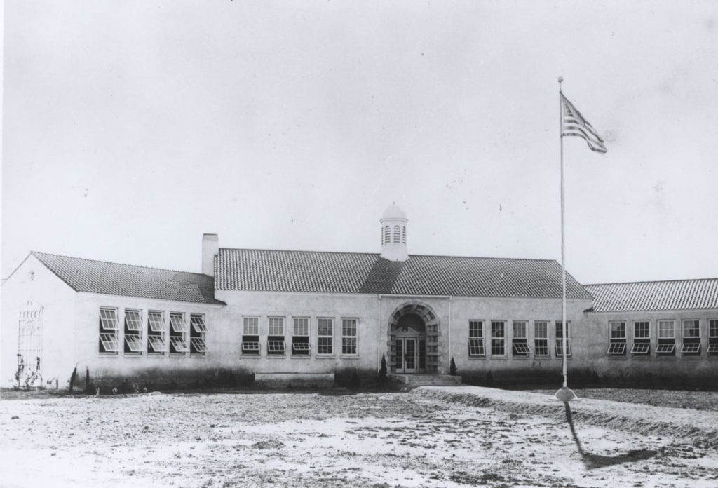 A vintage photograph of a tall one-story school building with a central wing with two groups of five windows and two flanking wings, each with five windows. The courtyard may be bare soil. A walkway to the front door contains a flagpole.