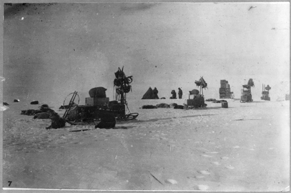 A vintage photograph of an antarctic scene of packed ice or snow. One tent, sledges with stacks of boxes, sled dogs, and a few human figures can be distinguished.