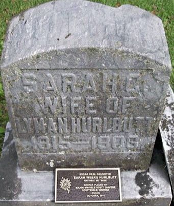 A snapshot of a gravestone with a small plaque attached. The gravestone reads, “Sarah G. Wife of Lyman Hurlbutt / 1815-1906.”